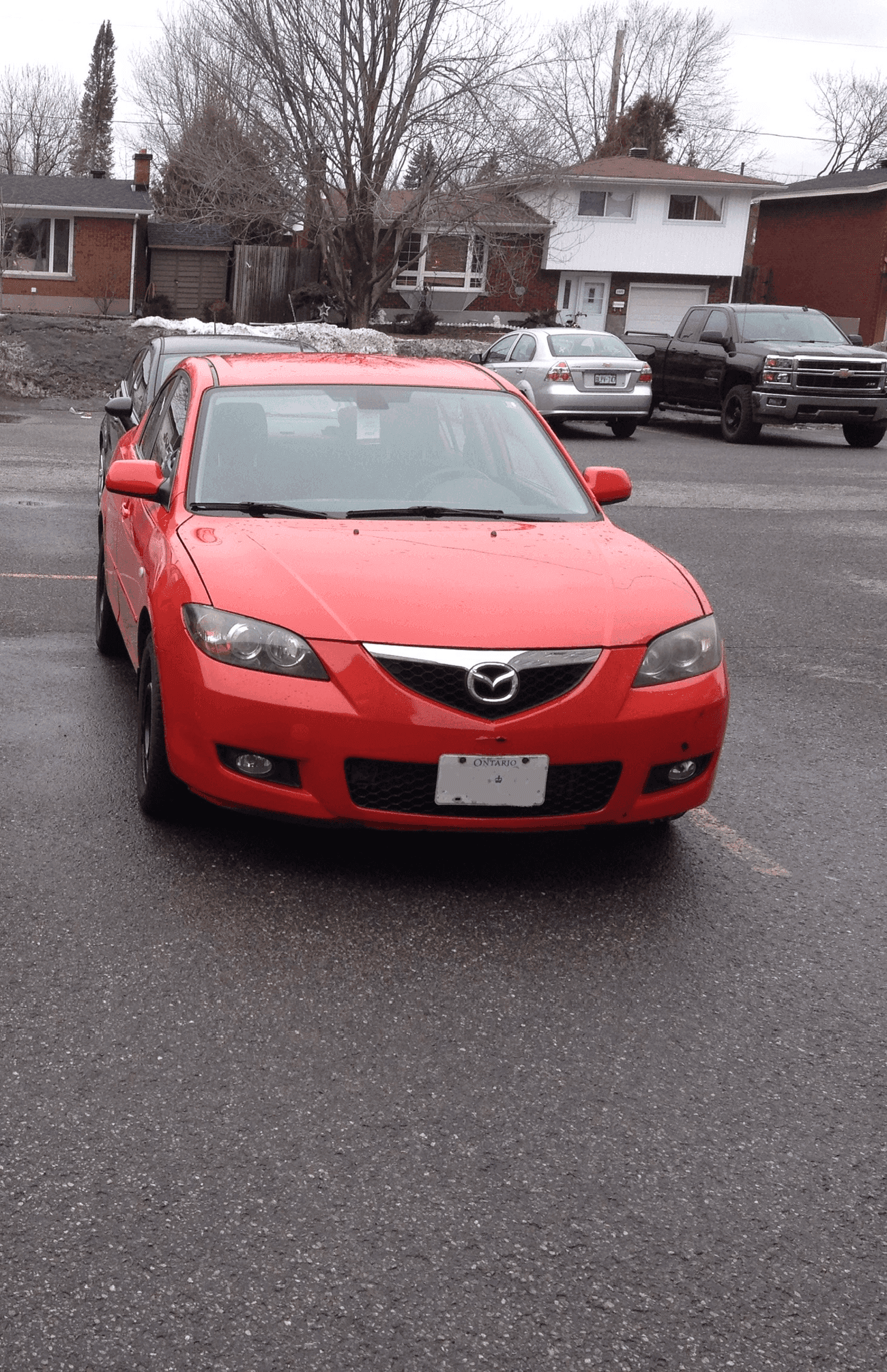 A red 2008 Mazda 3 photographed head-on in a residential parking area under a grey winter sky, bare trees and suburban houses behind it.
