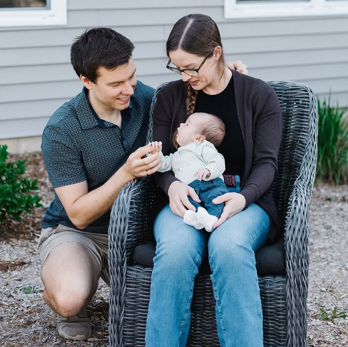 A couple outside their home with their baby: the mother sitting in a grey wicker chair holding the baby on her lap, the father crouched beside them holding the baby's hand, both smiling down at her.