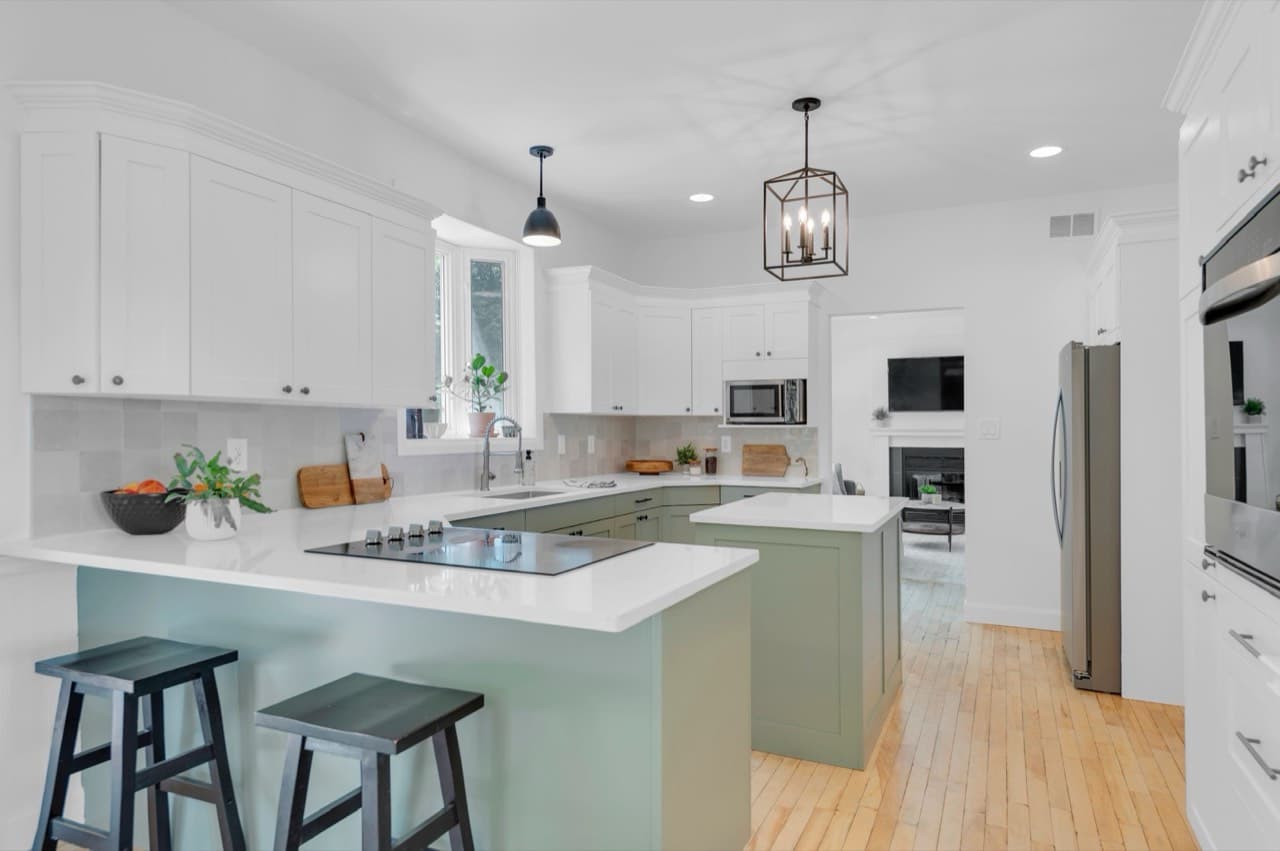 A renovated open kitchen with white and sage-green cabinetry, quartz counters, and pendant lighting.