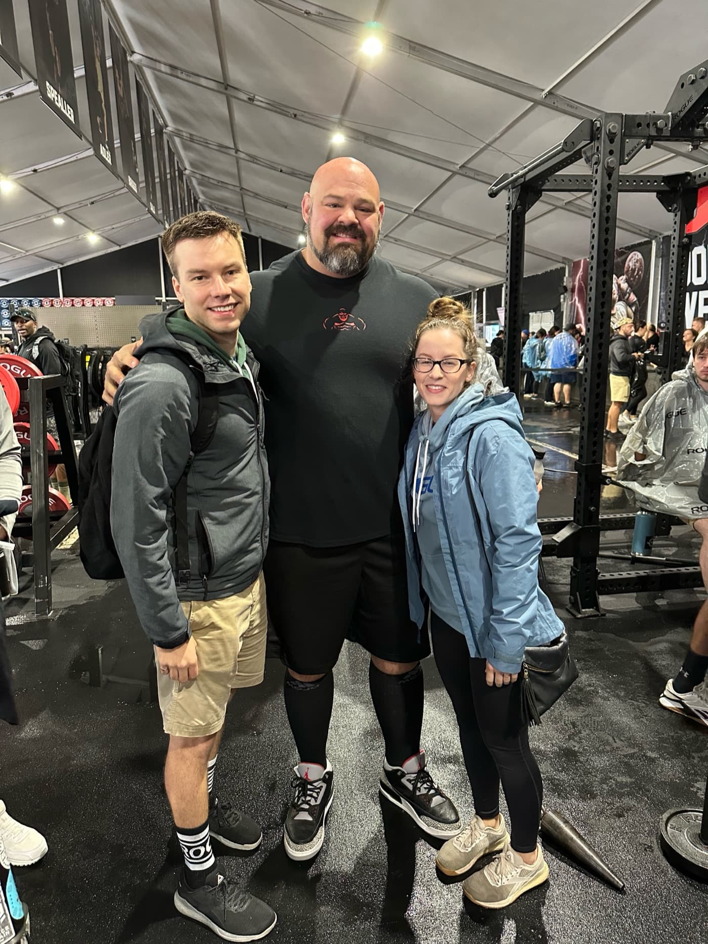 Three people posing arm in arm on a wet floor inside an equipment tent at a fitness competition, the very tall man in the middle towering over the couple on either side, weight racks and plates around them.