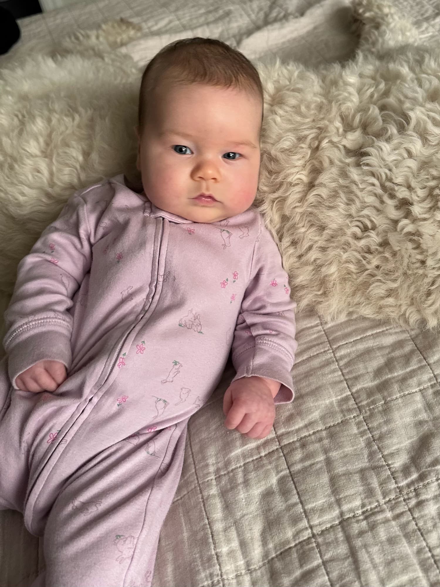 A baby in a pale pink bunny-print sleeper resting against Sasha, the family's fluffy white curly-coated dog, on a bed, looking at the camera.